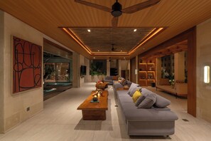 A modern living room with a wooden ceiling, a large gray sofa, a wooden coffee table, and a pool area visible through the glass wall.