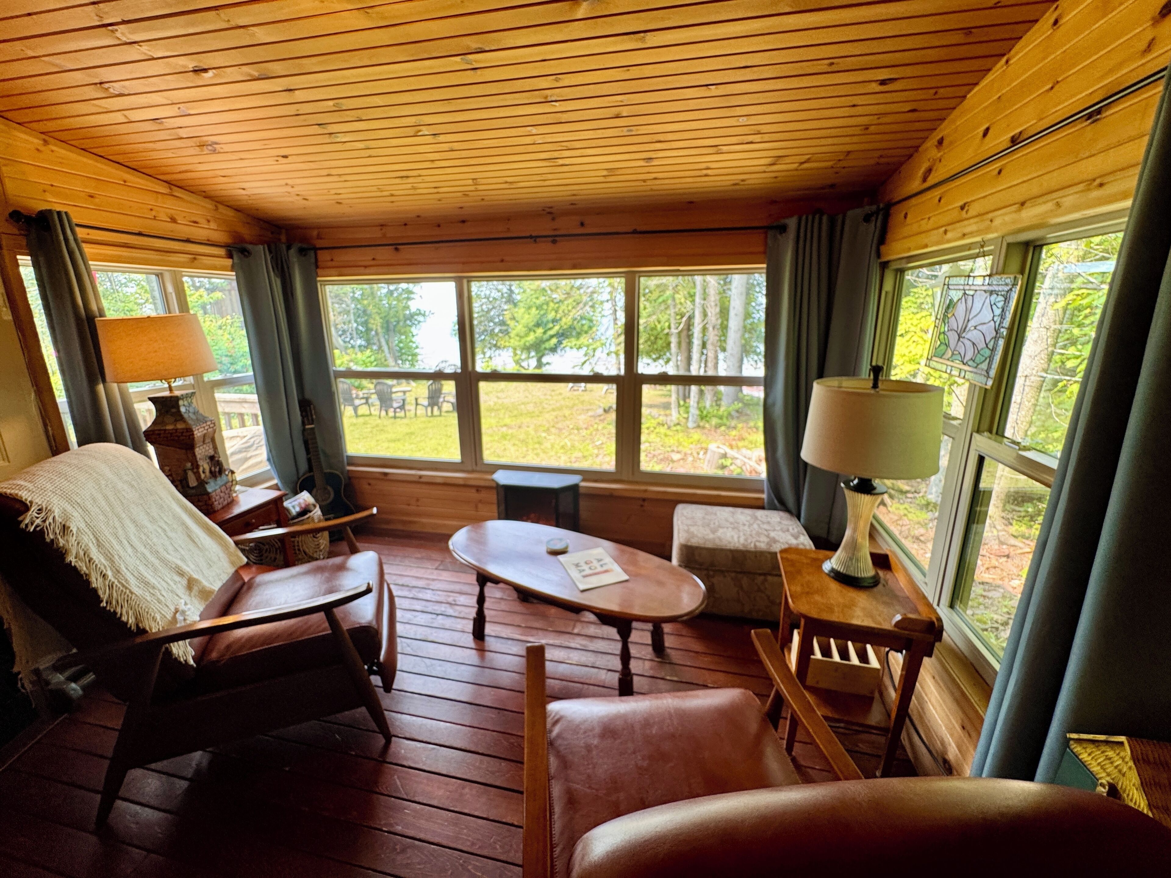 L’intérieur chaleureux d’un chalet avec un plancher en bois, une cheminée, un coin repas et de grandes fenêtres offrant une vue sur l’extérieur.