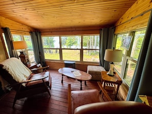 A cozy cabin interior with wooden floors, a fireplace, a dining area, and large windows offering a view of the outdoors.
