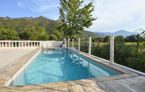 A rectangular outdoor swimming pool with a wooden deck, surrounded by a white railing, set against a backdrop of lush greenery and mountains.