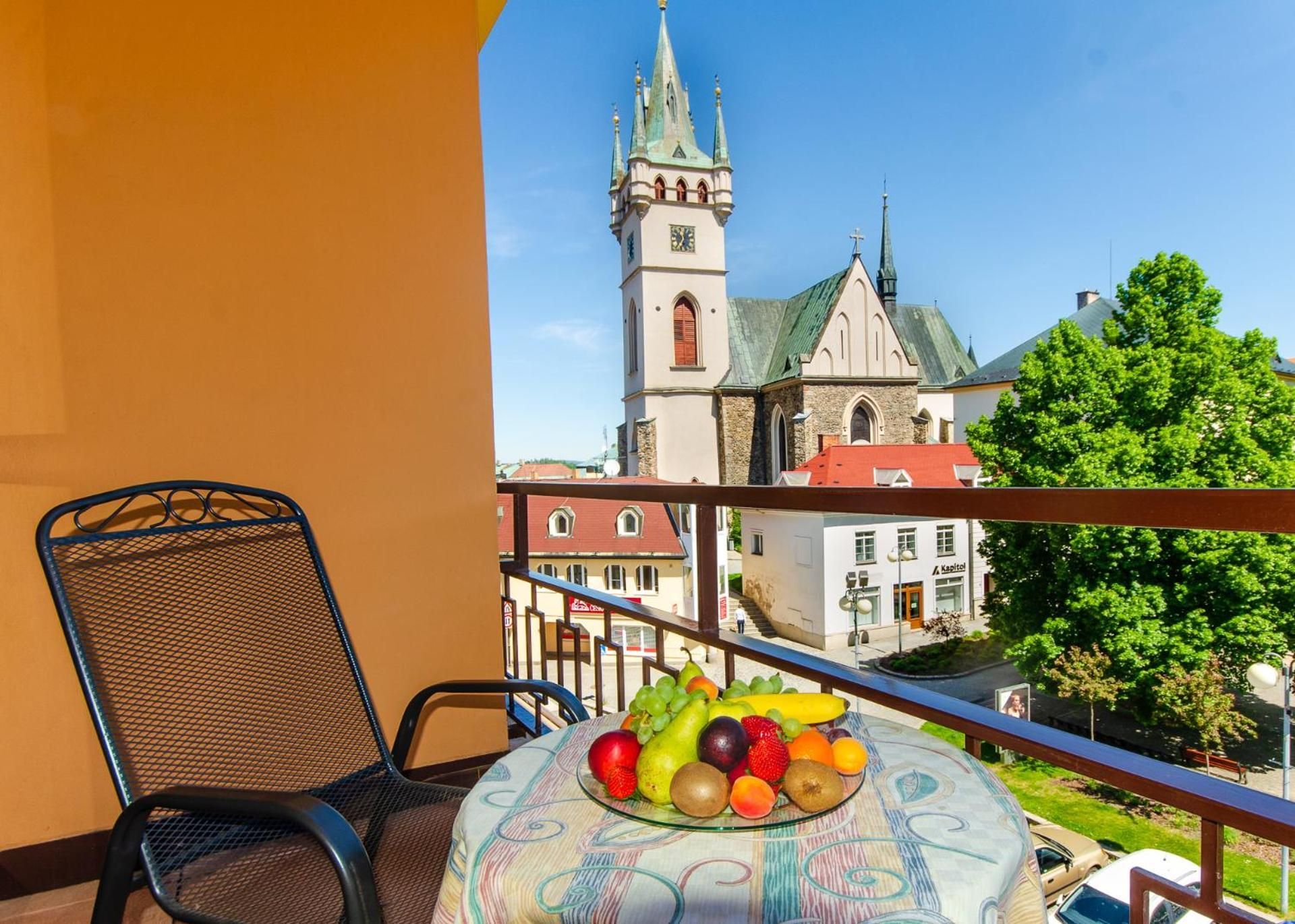 A balcony with a chair and a table with fruit, overlooking a church and buildings.