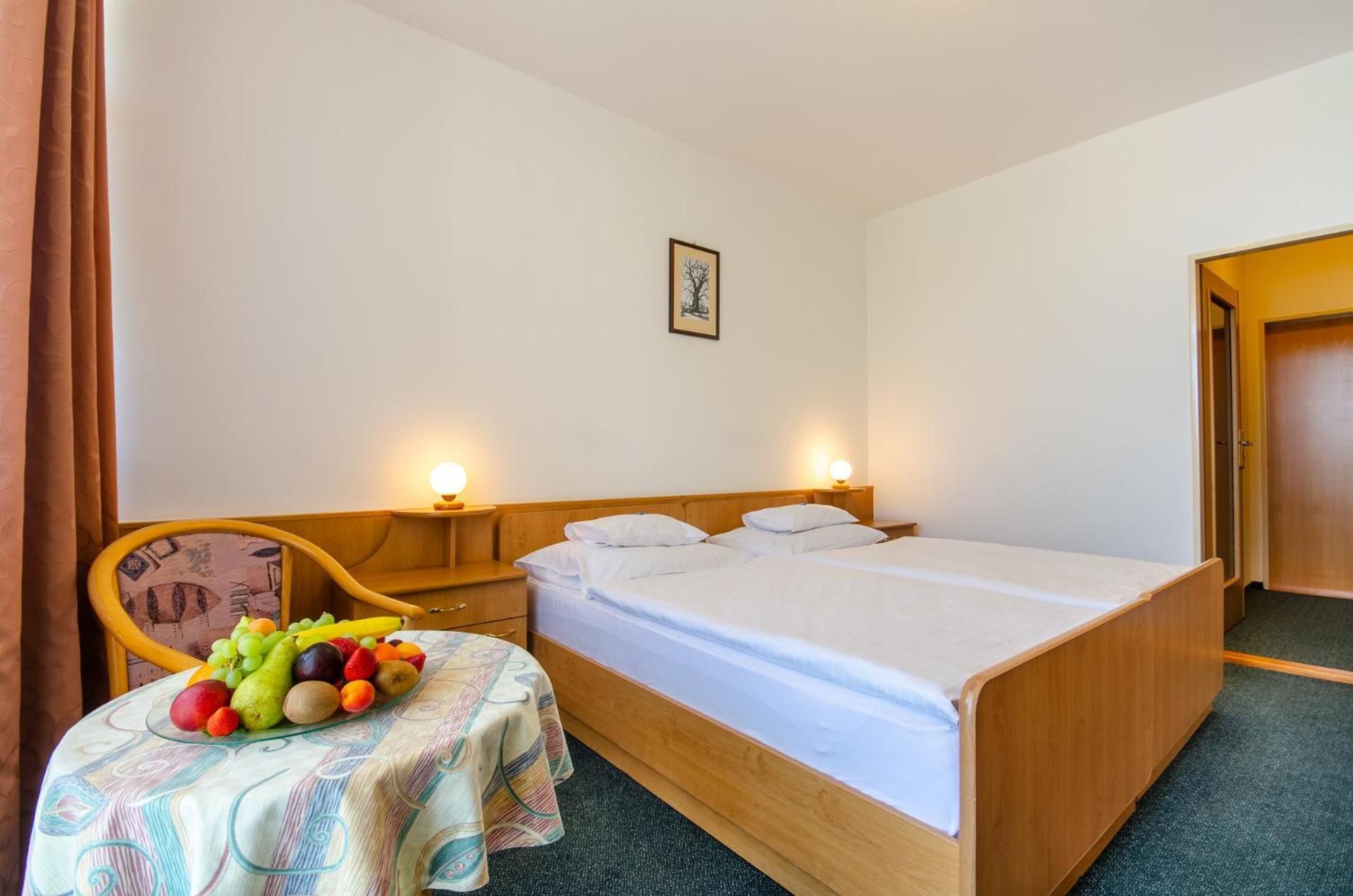A hotel room with a single bed, bedside tables, a chair, and a small table with fruit.