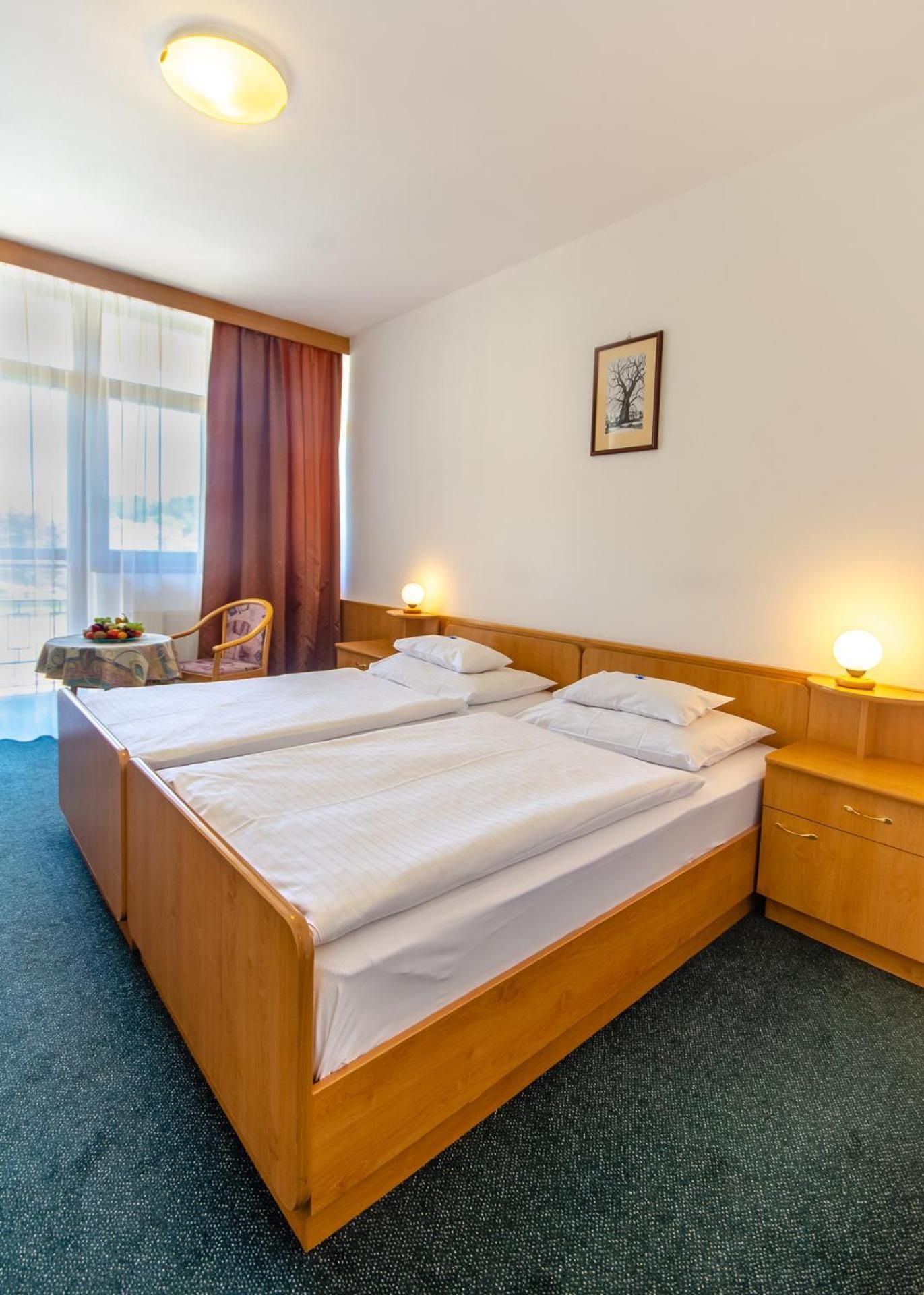 A double bed with wooden headboard and footboard, two bedside tables with lamps, a chair, a small table with fruit, and a framed picture on the wall.
