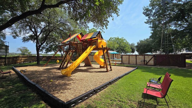 Ein Spielplatz mit Rutsche und Klettergerüsten, umgeben von einem Zaun und Bäumen.