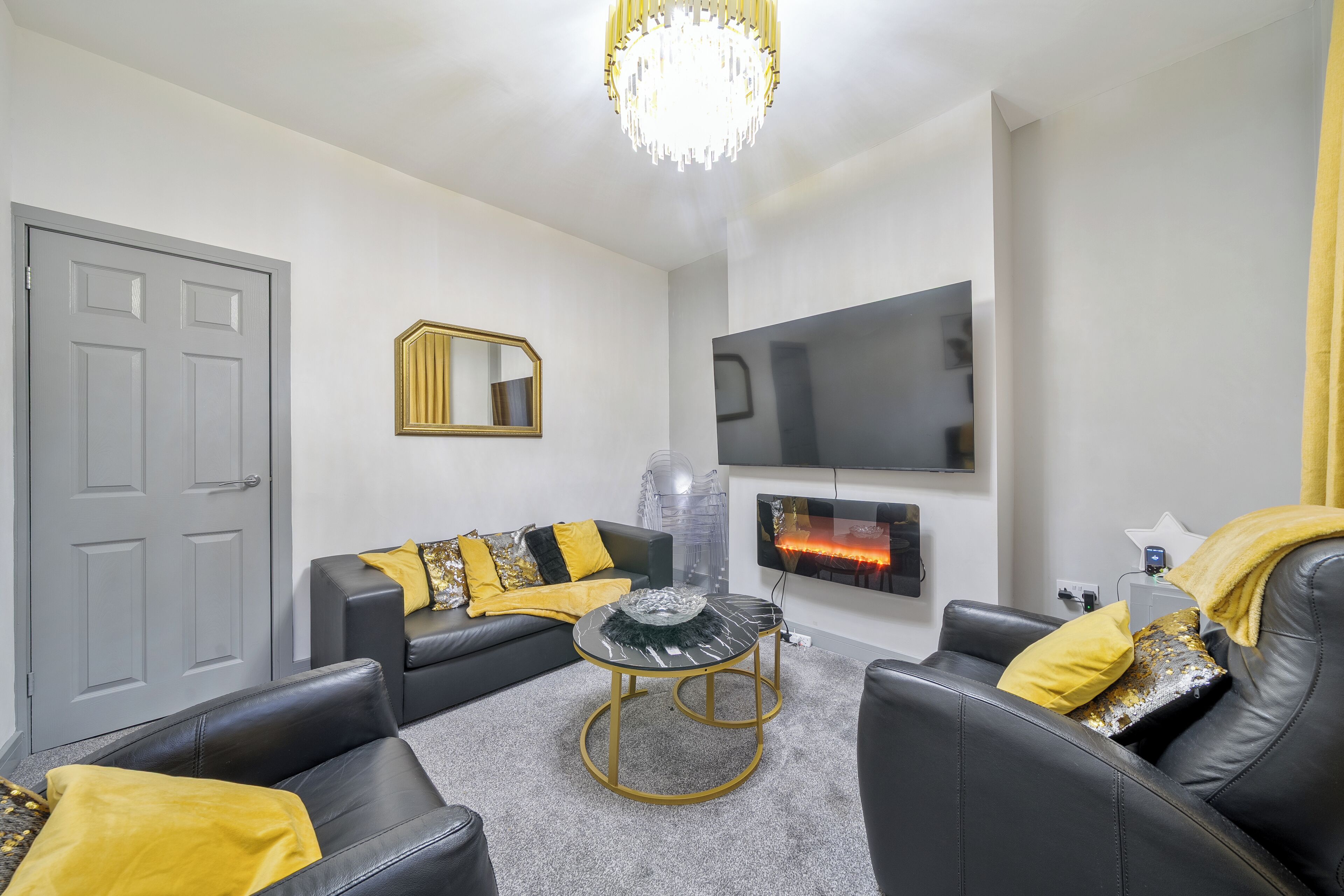 A modern living room with a grey door, a black leather sofa, a round coffee table, and a flat-screen TV mounted on the wall.