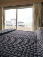 Una cama con cubierta acolchada, una ventana con cortinas y vistas a edificios y una playa.