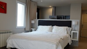 Habitación de hotel moderna con una cama grande, mesitas de noche, una ventana y vista al área de la cocina.