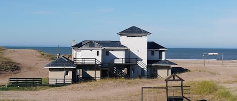 A coastal building with multiple levels and a prominent tower, situated on a sandy beach with a clear view of the ocean.
