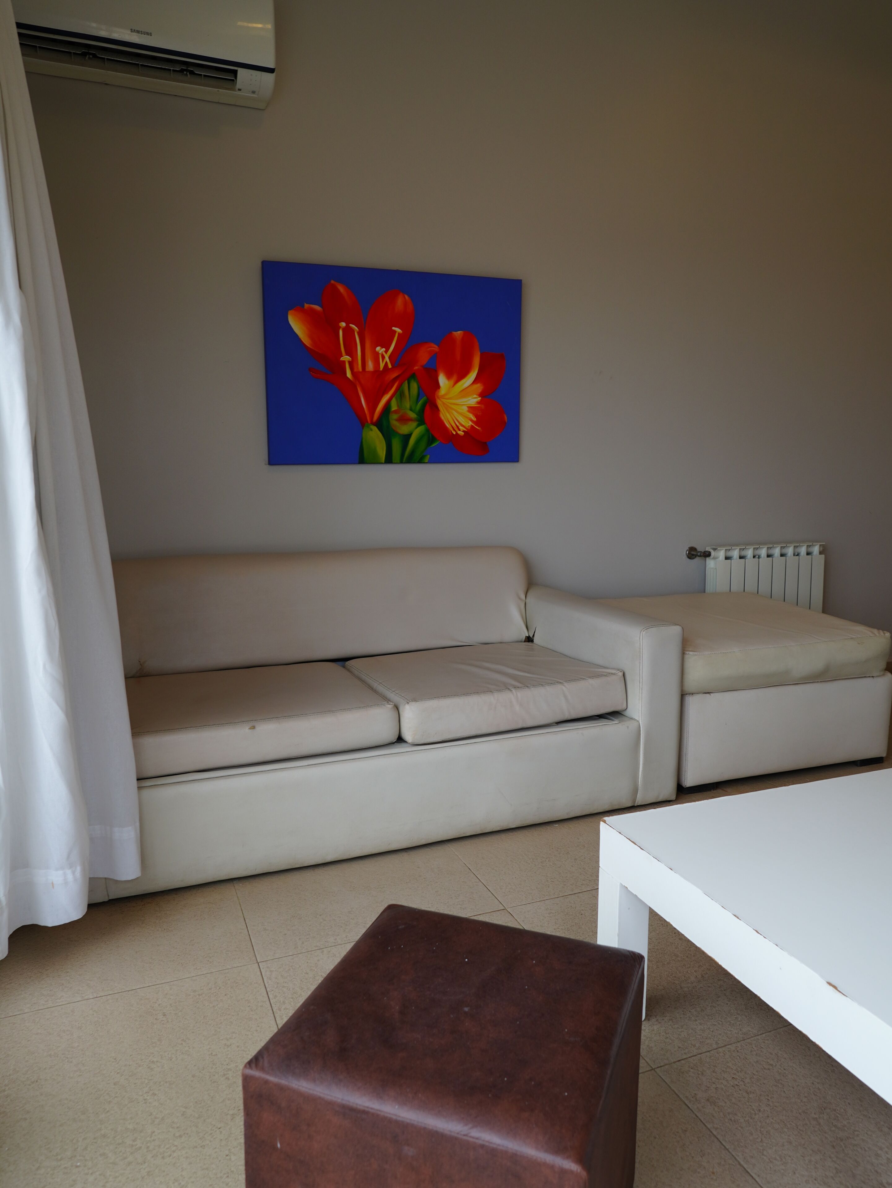 Un salon avec un canapé beige, un pouf marron, une table basse blanche et un tableau représentant des fleurs rouges accroché au mur.
