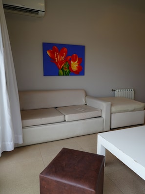 A living room with a beige sofa, a brown ottoman, a white coffee table, and a painting of red flowers on the wall.