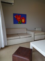 Una sala de estar con un sofá color beige, un reposapiés marrón, una mesa de centro blanca y un cuadro con flores rojas en la pared.