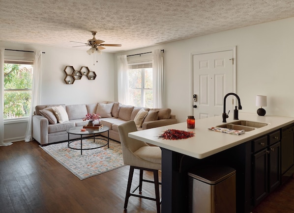 A modern kitchen with a central island, a dining area with a sofa and coffee table, and a ceiling fan.