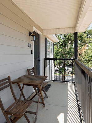 A covered porch with a wooden table and two chairs, a black metal railing, and a door with the number 201.