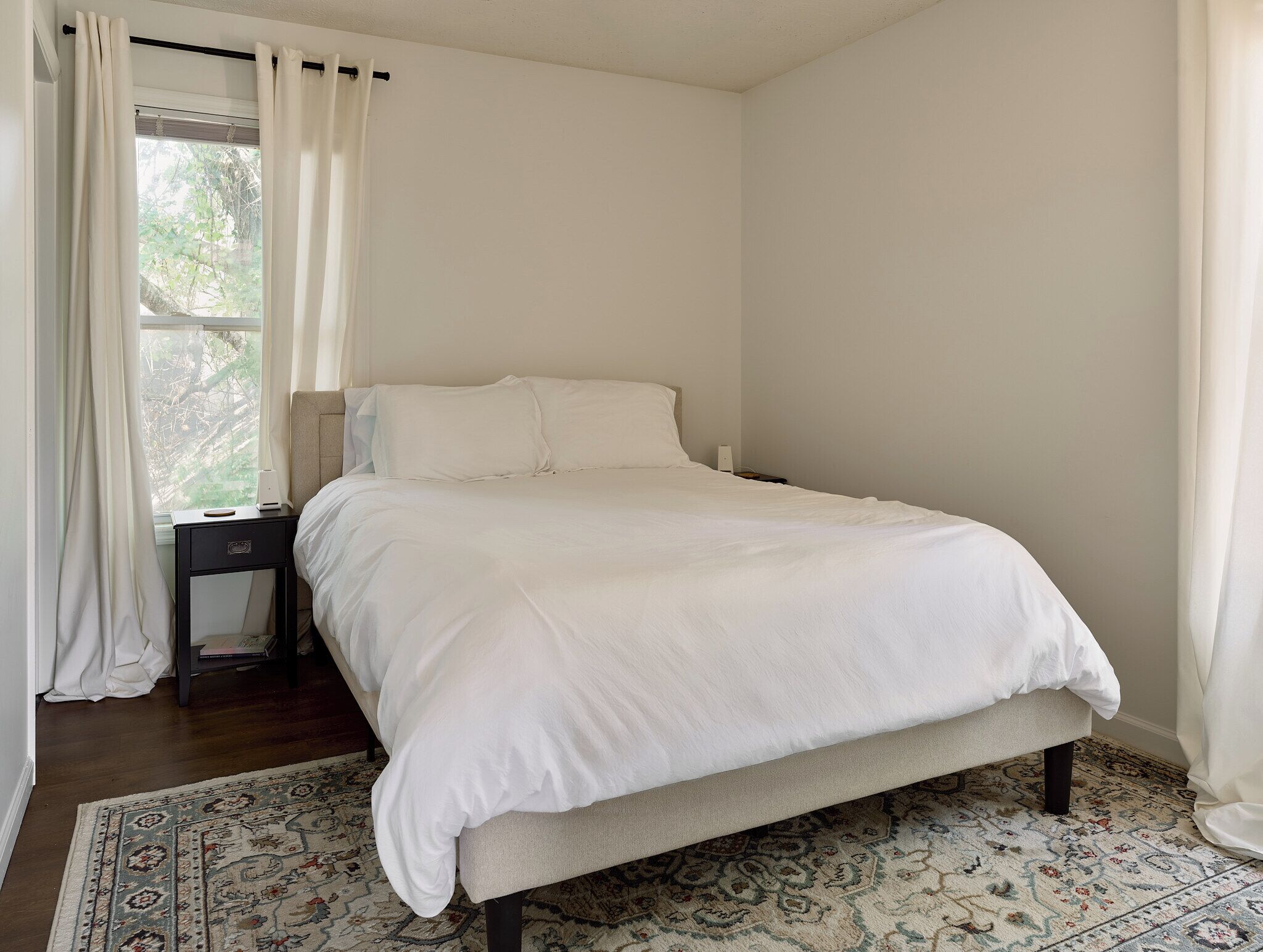 Un dormitorio con cama, mesita de noche, ventana con cortinas y una alfombra estampada.