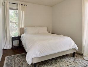 Un dormitorio con cama, mesita de noche, ventana con cortinas y una alfombra estampada.