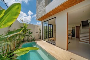 Ein moderner Poolbereich im Freien mit einer Steinmauer, Glastüren zu einem Wohnbereich und üppiger tropischer Bepflanzung.