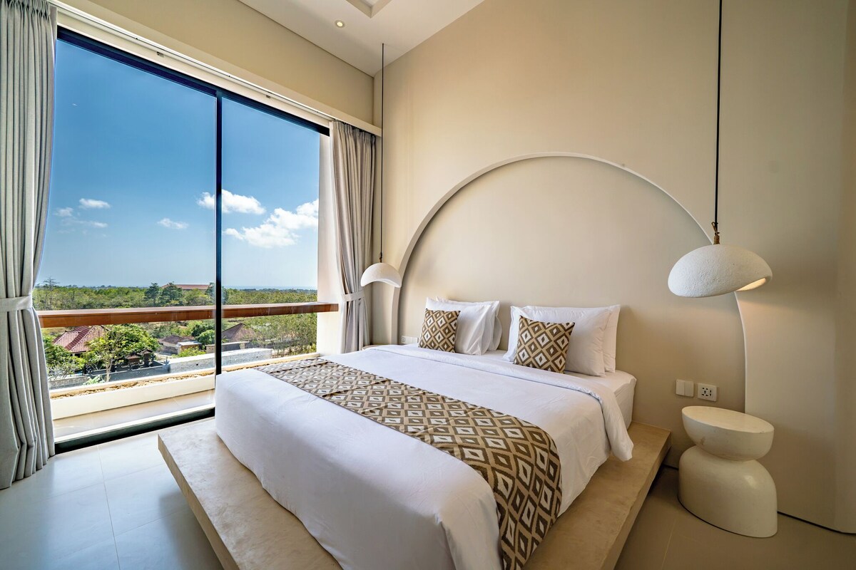 Ein modernes Schlafzimmer mit einem großen Bett, zwei Nachttischlampen, Blick auf die Landschaft und einer geschwungenen Wandgestaltung.