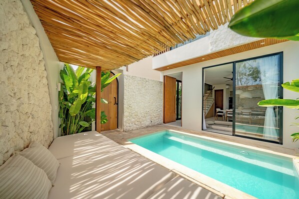 Un área de piscina moderna al aire libre con un muro de piedra, una pérgola de madera y un ventanal que da a un patio cubierto.