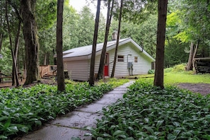 A cabin surrounded by dense forest, with a wooden deck and a pathway leading to the entrance.