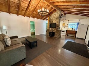 A living room with a stone fireplace, wooden beams, a sofa, and a television. - Hidden River Retreat | Secluded Riverfront Cottage (Markdale)