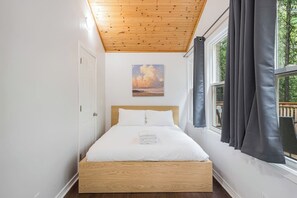 A single bed with a wooden headboard in a room with a wooden ceiling, a window with curtains, and a painting on the wall. - Hidden River Retreat | Secluded Riverfront Cottage (Markdale)