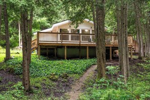 A two-story wooden cabin with a deck, surrounded by dense forest.