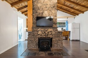 A living room with a stone fireplace, a television mounted on the fireplace, and a kitchen area in the background. - Hidden River Retreat | Secluded Riverfront Cottage (Markdale)