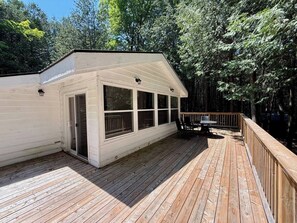 A covered outdoor deck with a wooden floor, a white building with large windows, and a view of a forested area.