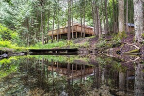 Une cabane dotée d’une terrasse qui surplombe un étang, le tout étant entouré d’une forêt dense.