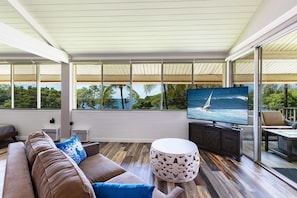 A modern living room with a large TV, comfortable seating, and a view of the ocean. - Ocean Front Vibes Maui (Kihei)