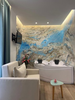 A spa room with a marble wall, a white bathtub, a beige sofa, and a small table with a pink object. - Hotel Ilio (Ksamil)