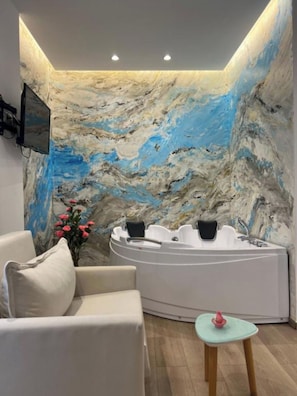 Ein Spa-Raum mit Marmorwand, einer weißen Badewanne, einem beigefarbenen Sofa und einem kleinen Tisch mit einem pinkfarbenen Gegenstand.