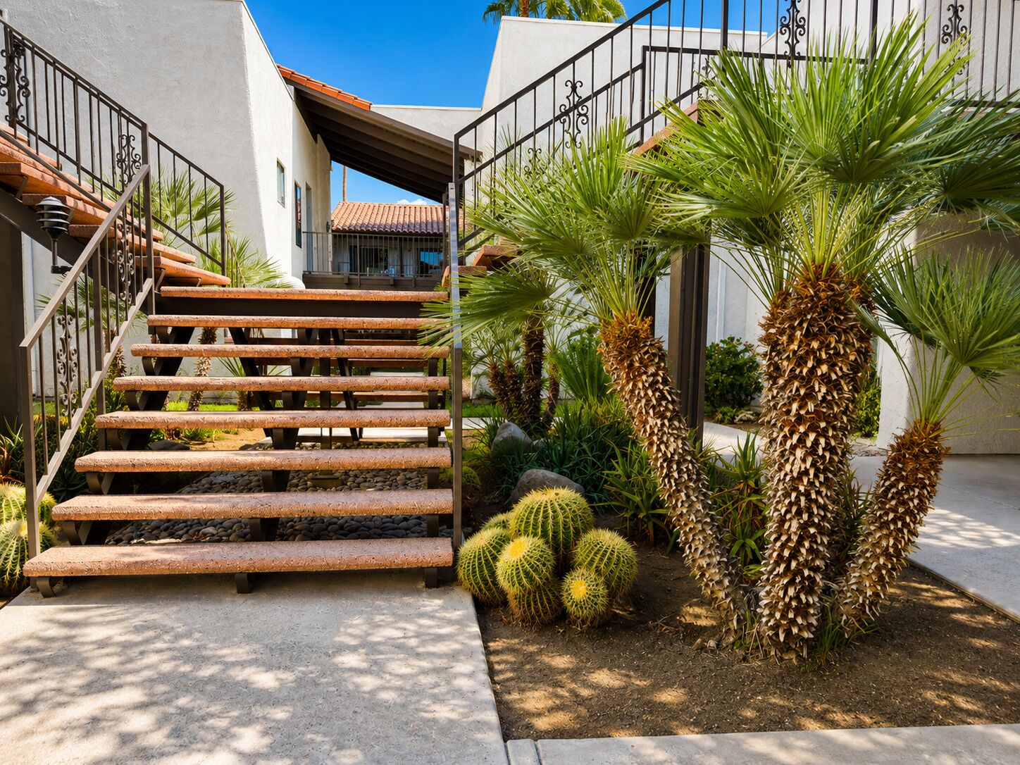 A staircase with a metal railing leading to a covered area, surrounded by various plants including palm trees and cacti.