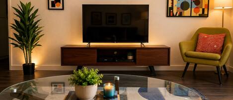 A modern living room with a glass coffee table, a flat-screen TV, a potted plant, a bookshelf, and a yellow armchair.