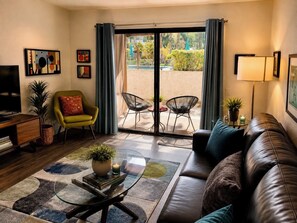 A living room with a sofa, armchairs, a coffee table, a TV, and a sliding glass door leading to an outdoor patio with chairs and a table. - Modern  1-bedroom condo with AC in sunny Palm Springs (Palm Springs)