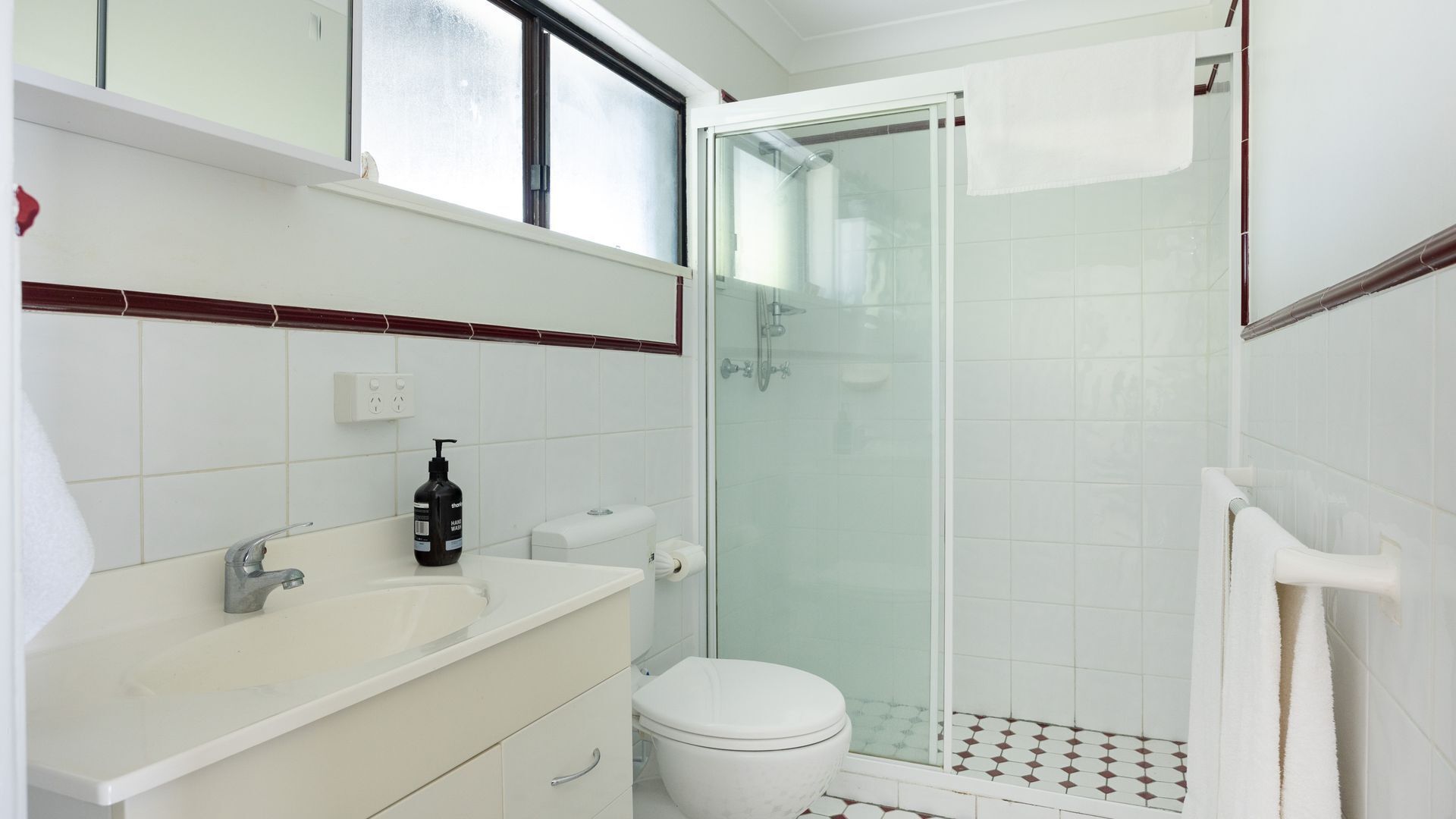 A bathroom with a glass-enclosed shower, a white toilet, a sink with a bottle of shampoo, and a towel rack.