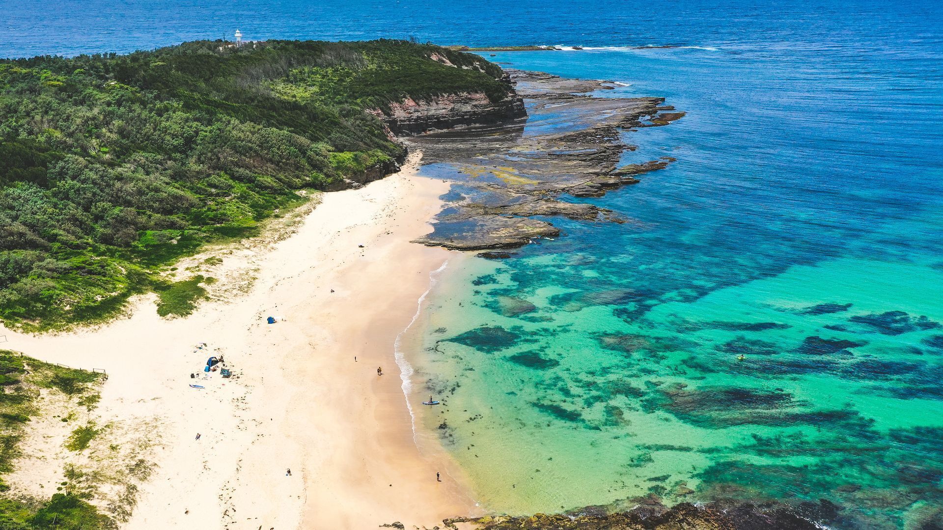 An aerial view of a beach with clear turquoise waters, a sandy shore, and a rocky outcrop with vegetation.