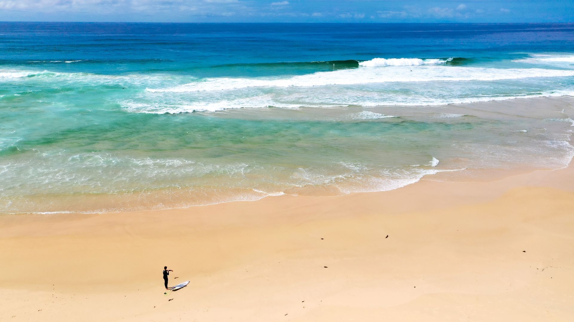 Een strand met helder turquoise water, een zandstrand en een persoon met een surfboard.