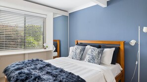A bedroom with a bed, a window with blinds, a bedside lamp, and a blue wall. - Rennies Sands | A Surfer's Dream by Rennies Beach (Ulladulla)