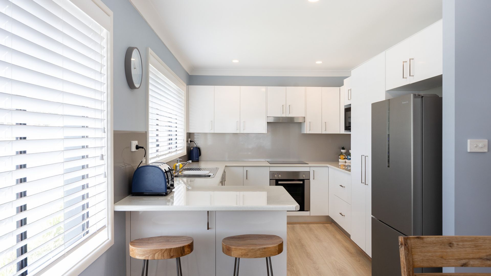 A modern kitchen with white cabinetry, a stainless steel refrigerator, and a built-in oven. There are two wooden stools and a window with blinds.