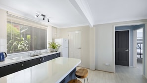 A modern kitchen with a central island, a built-in sink, and a refrigerator. There is a window with blinds, a door leading to another room, and a staircase visible in the background. - Rennies Sands | A Surfer's Dream by Rennies Beach (Ulladulla)