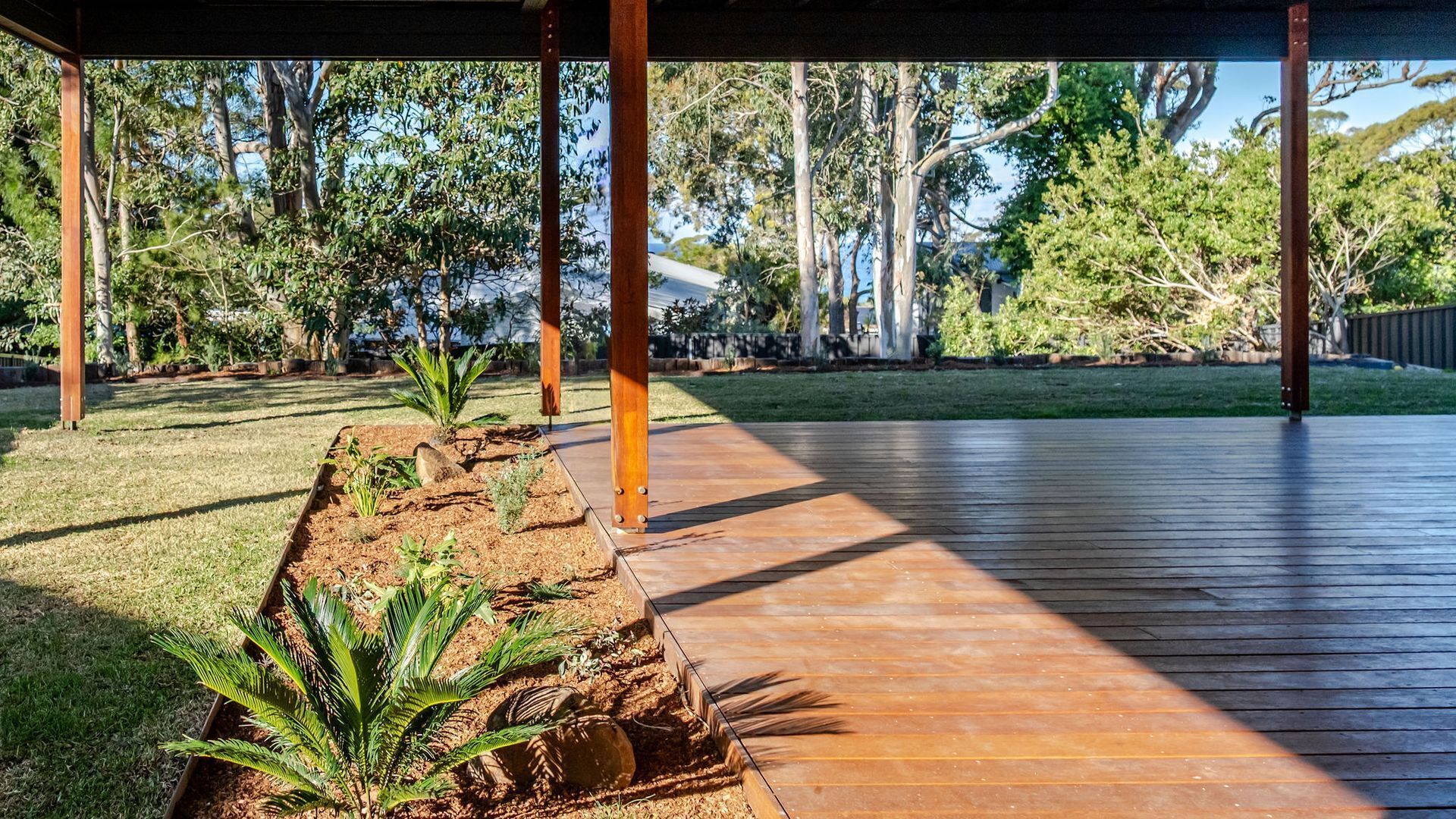 Een overdekt buitengebied met een houten terras, omgeven door een tuin met diverse planten en bomen.
