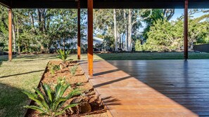 A covered outdoor area with a wooden deck, surrounded by a garden with various plants and trees. - Molly-a-la-Plage - Beautiful, spacious and modern beach house in Mollymook Beach (Mollymook Beach)