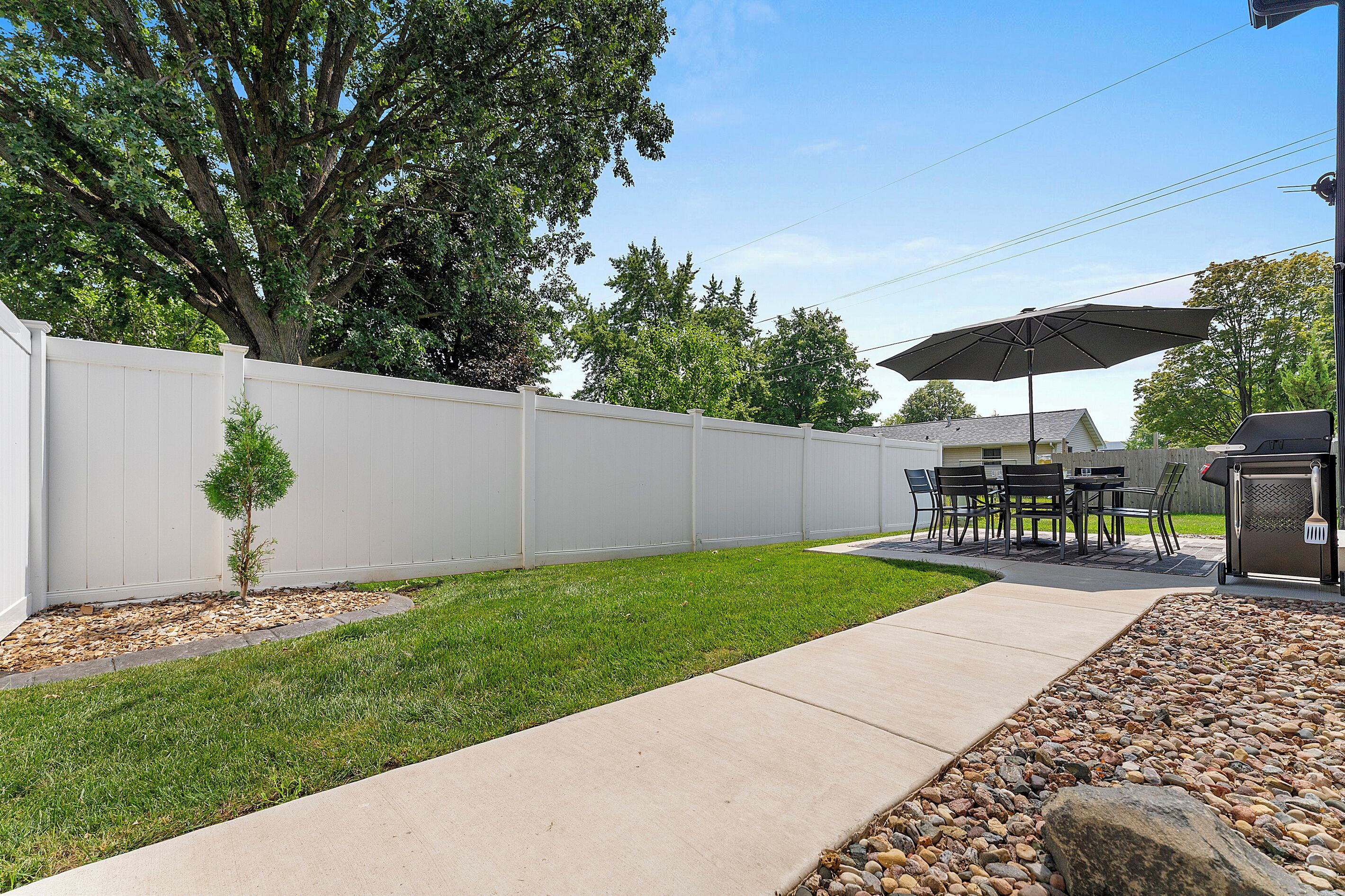 A backyard with a white fence, a patio with a table and chairs, and a grill.