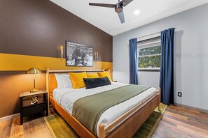 A bedroom with a bed, nightstand, lamp, ceiling fan, window with curtains, and a wall-mounted picture.