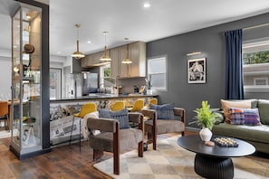 Een moderne woonkamer met een bank, stoelen, een salontafel en een keukenhoek met kastjes en een koelkast.