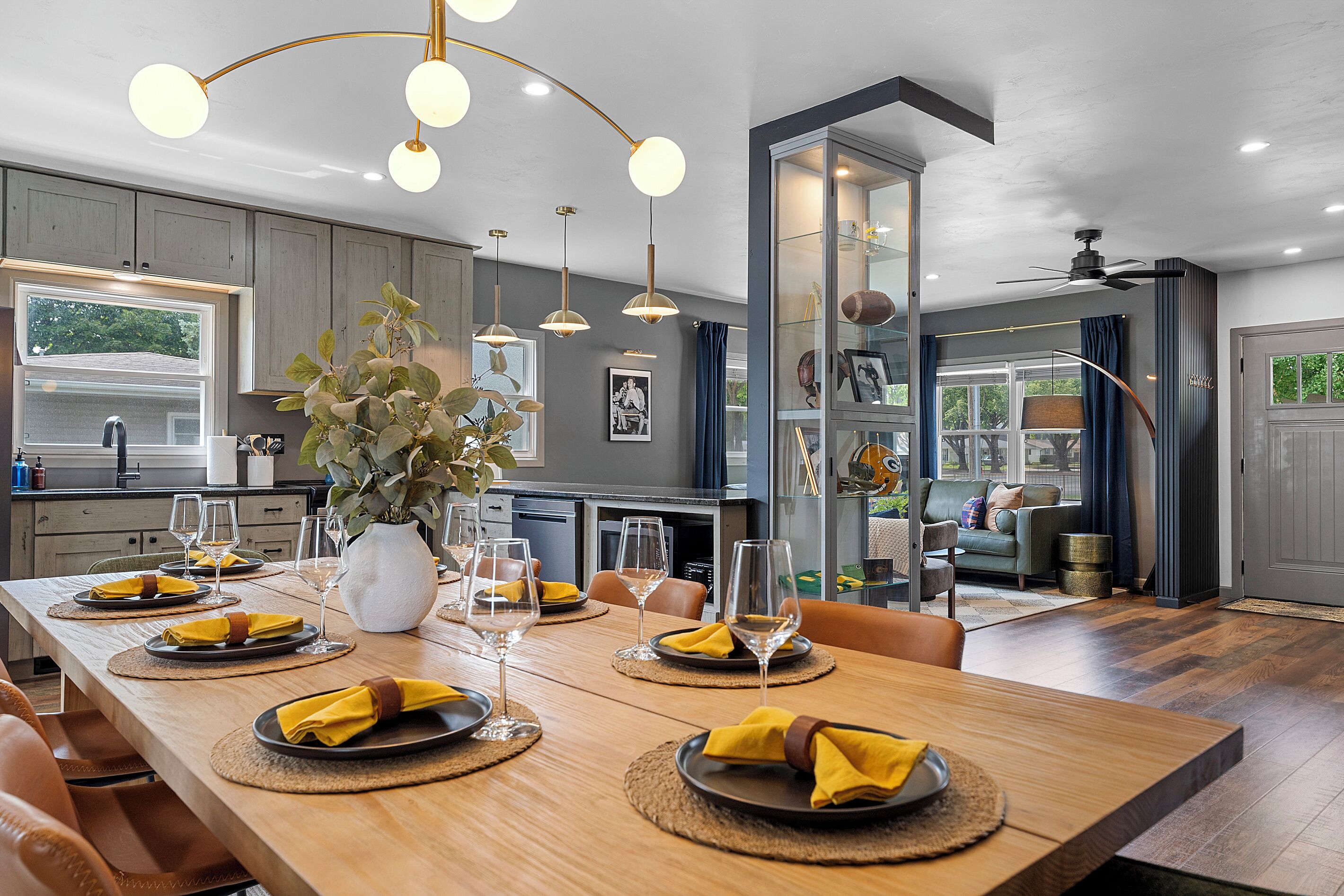 Un comedor moderno con una mesa de madera dispuesta para una comida, un florero con plantas y luminarias colgantes.