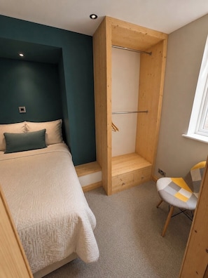 A bedroom with a bed, a wooden wardrobe, a chair, and a window. - Apartment Sleeps 4 - Town Centre & Near Beach (Newquay)