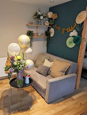 Ein Zimmer mit einem grauen Sofa, einem kleinen runden Tisch mit Blumen in einer Vase und einer Wand mit einer 'Happy Birthday'-Dekoration.