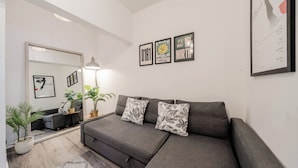 Ein modernes Wohnzimmer mit einem grauen Sofa, zwei dekorativen Kissen, einem kleinen Beistelltisch, einer Stehlampe und einem Spiegel. An der Wand hängen gerahmte Bilder und es gibt zwei Zimmerpflanzen.